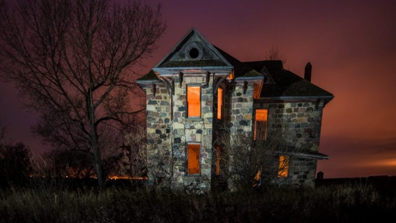 An abandoned stone house with orange-lit windows on a cloudy night.