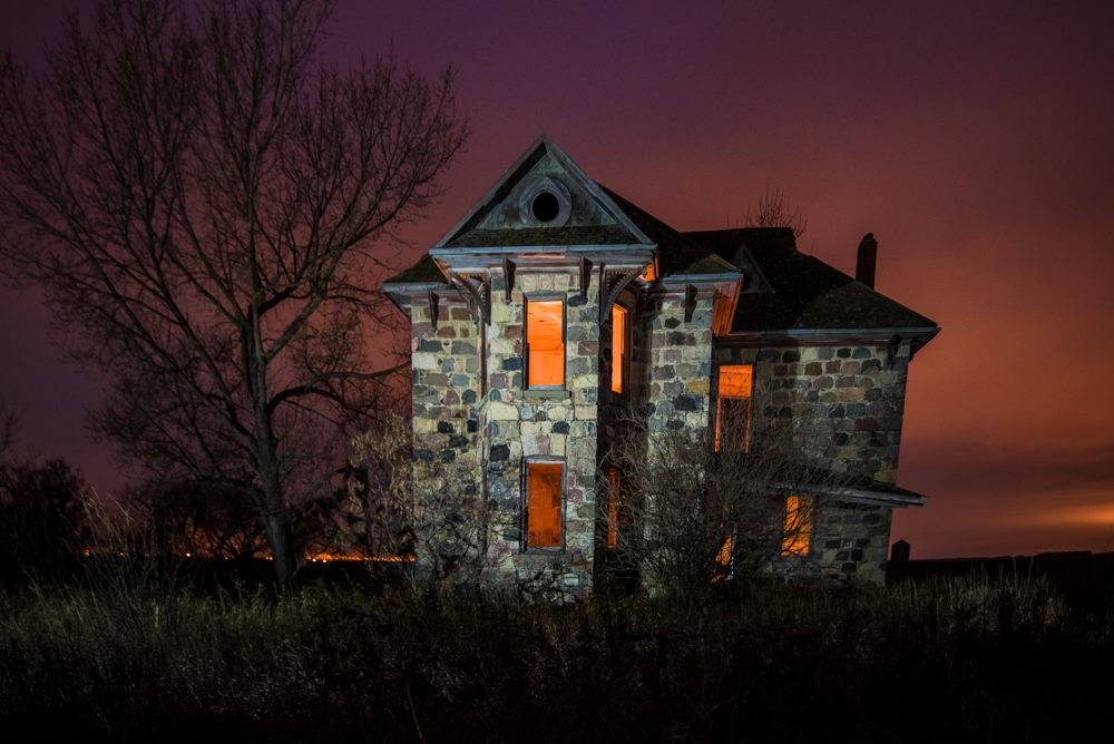 An abandoned stone house with orange-lit windows on a cloudy night.