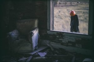 A pumpkin-head figure lurks outside the glassless window of an abandoned house.