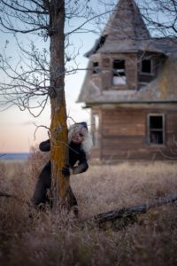 A creepy doll peeking out from behind a leafless treek with an abandoned wooden house in the background.