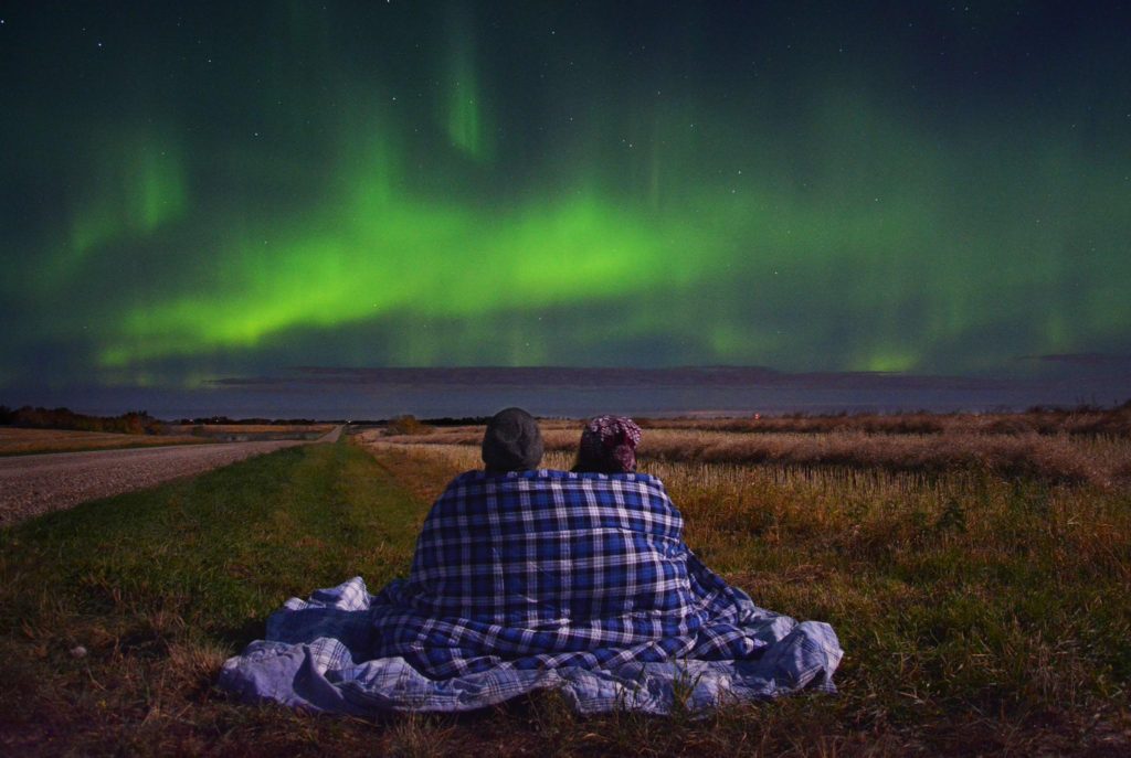 Photo of a couple cozying up together under a blanket while watching the northern lights dance over an open field.