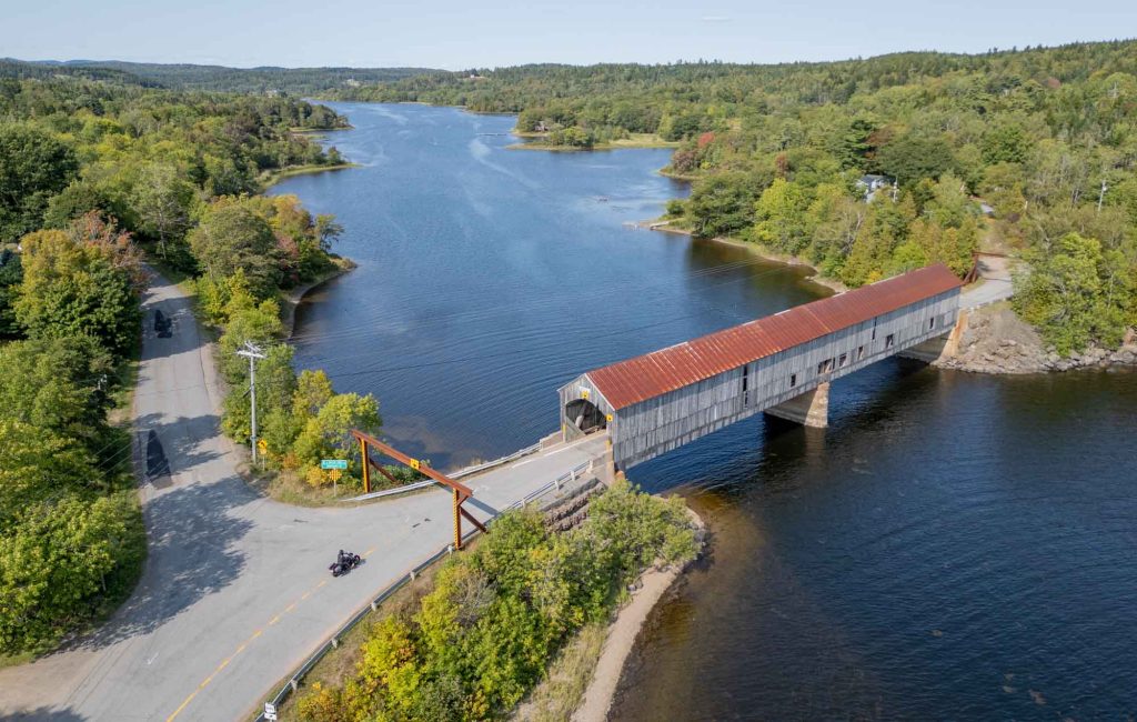 Red-roofed covered bridge over a river with a motorcycle approaching the entry to the bridge.
