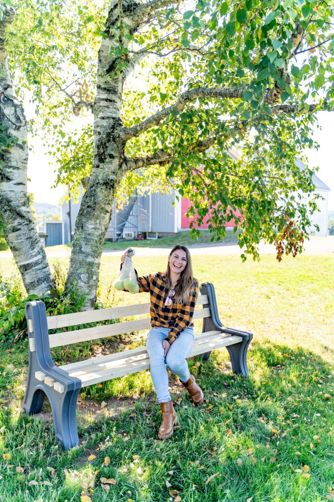 A woman in a yellow plaid shirt sitting on a wooden bench under a tree, holding up a bag of apples. 