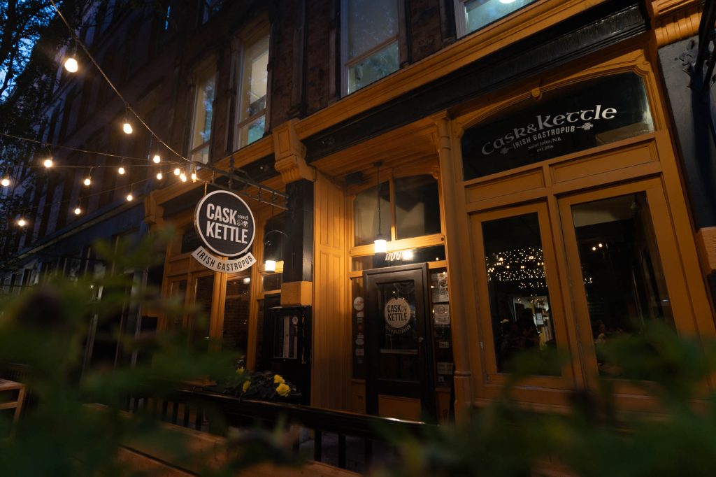 A nighttime view through greenery of the lit up sign of the Cask and Kettle gastropub and it's front door.