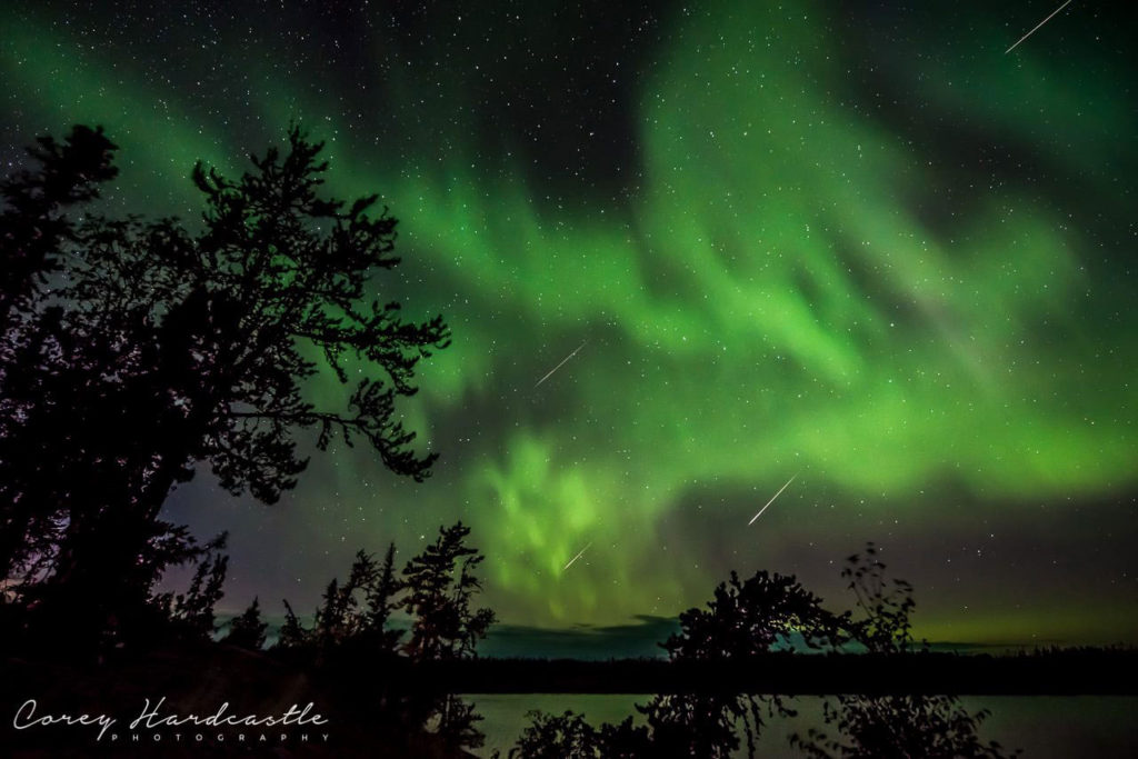 Green northern lights dancing over a lake with three meteors flashing through the sky.