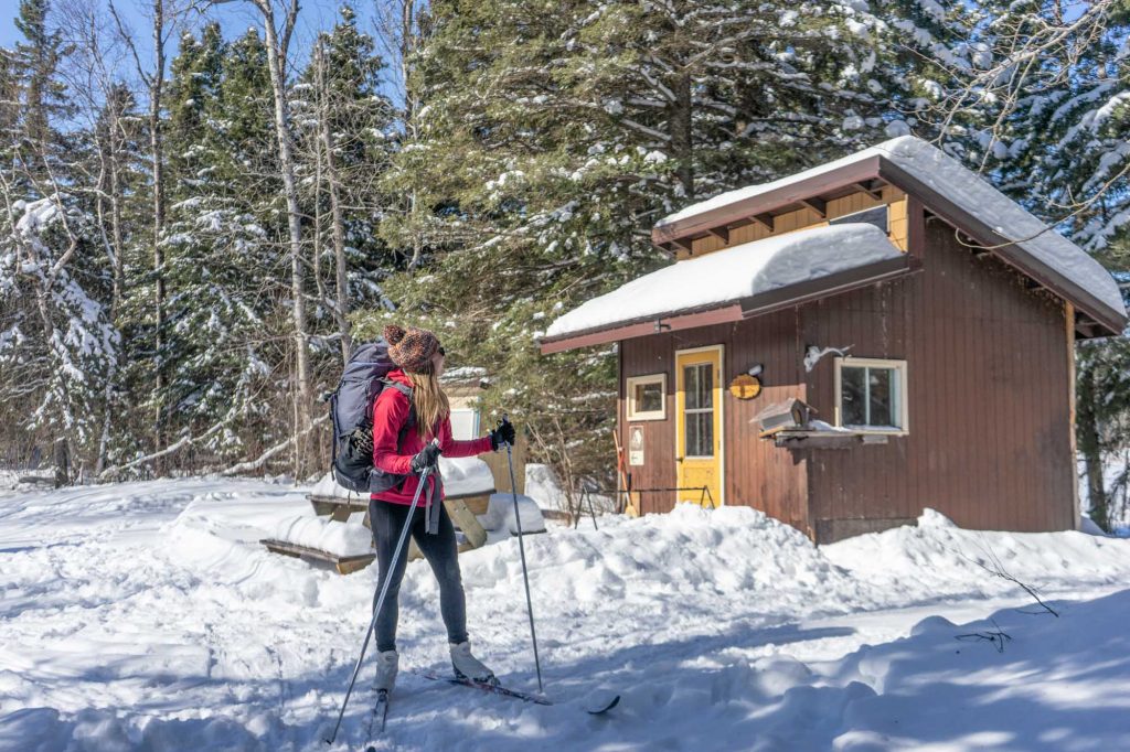 Girl in cross-country skis standing looking back at a small ski shelter in the snow
