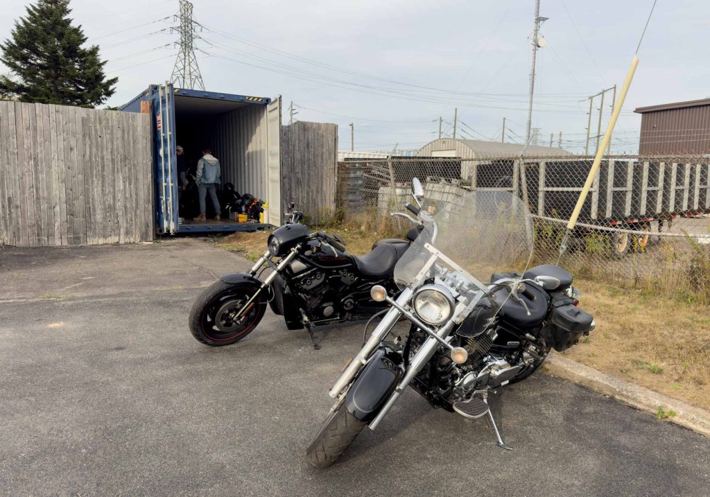 Two motorcycles sit in front of a sea can in a parking lot.