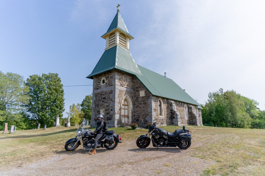 Two motocycles, one with a woman on it, in front of a green-roofed church with a tall steeple. The church is made out of stone. 