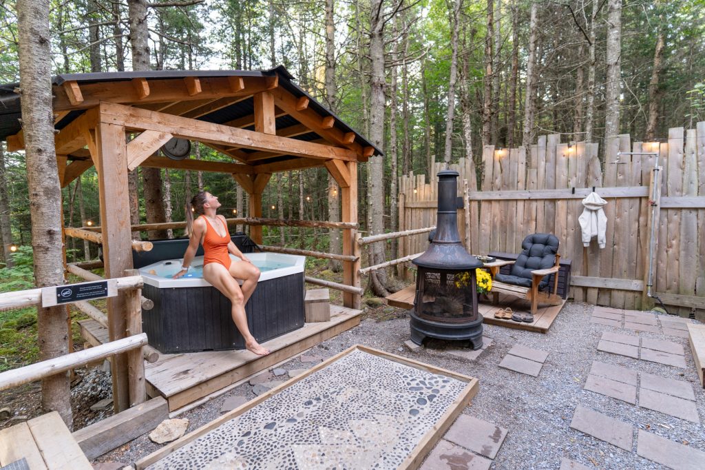 A woman leaning on a hot tub under a framed wooden roof with an outdoor firepit and chair next to it.
