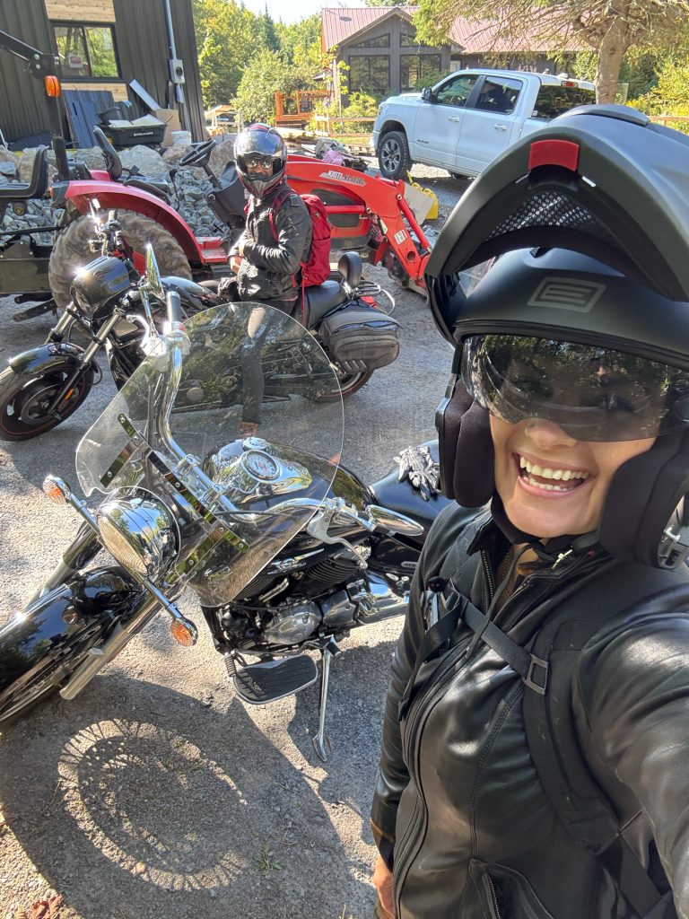 A selfie of a woman in a helmet on a motorcycle with a man on a motorcycle in the background.