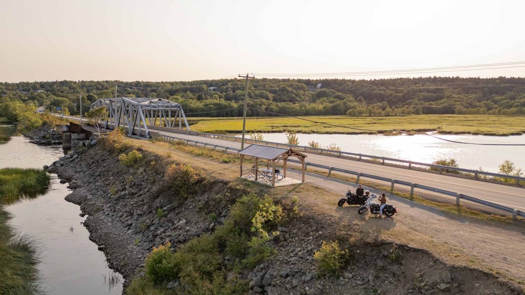 A man and woman each on motorcycles parked on the side of a highway with a river running under a bridge in the background.