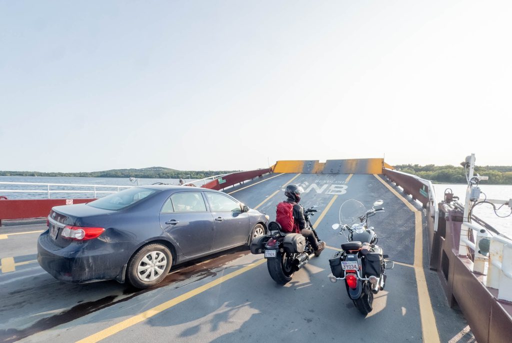 A car and two motocycles, one with a man on it, sit side-by-side at the front of a vehicle ferry crossing a river.