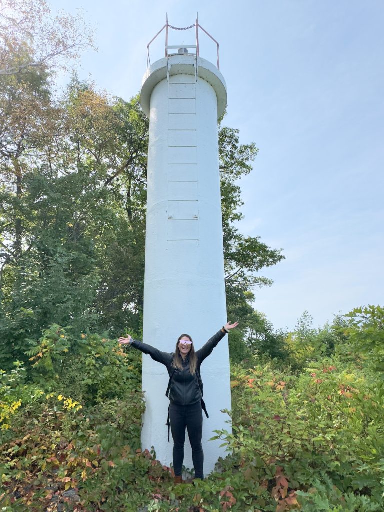 A woman standing with arms up in front of a lighthouse that it a circular pole.