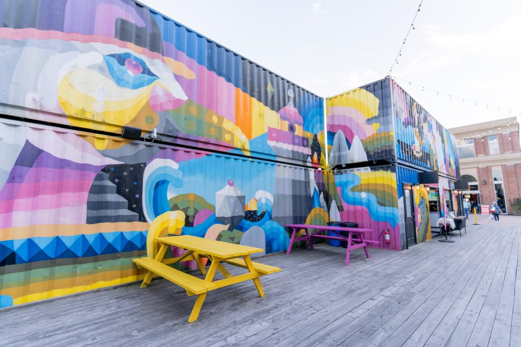 Two levels of colourfully painted  sea cans stacked on top of each with with yellow and purple picnic tables in front.