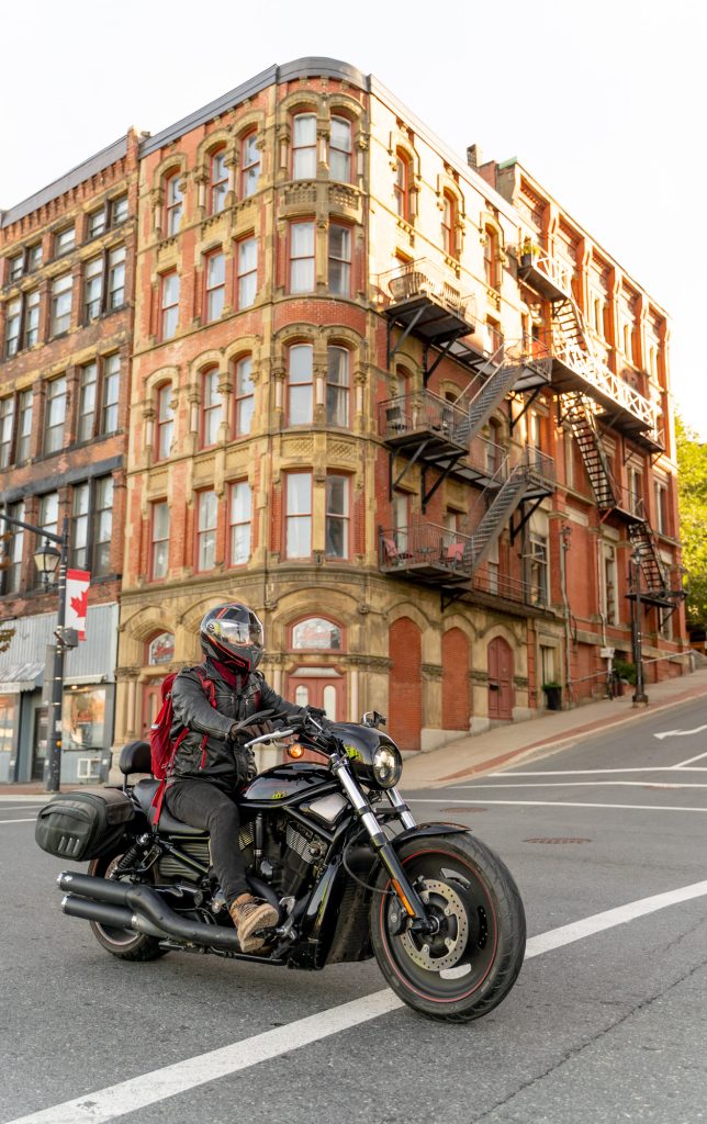 A man riding past on a motorcycle with a red and brown building with exterior staircases in the background.