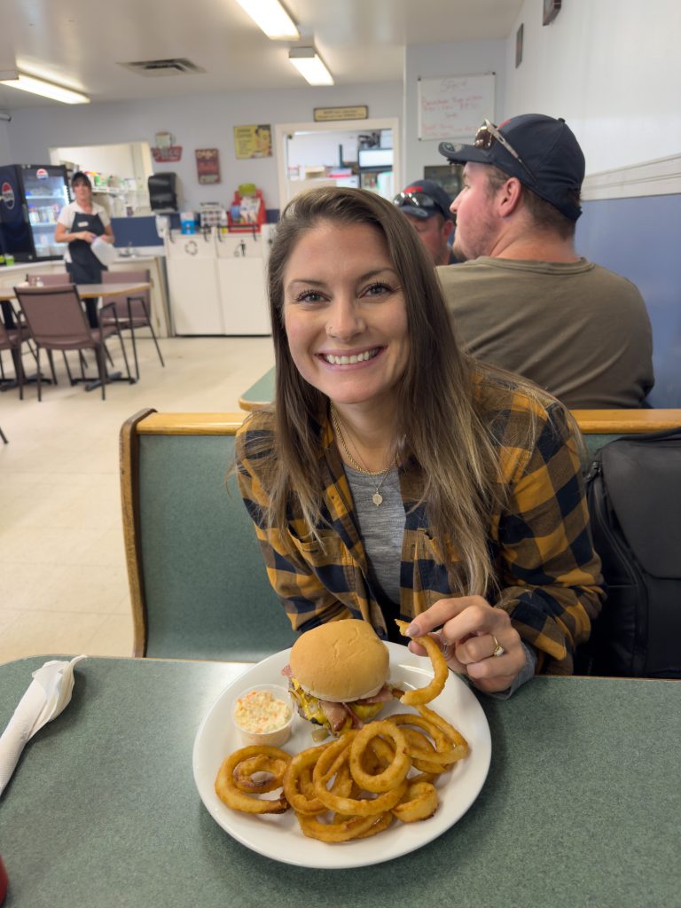 A woman with a burger and onion rings on a plate in a booth in a diner.