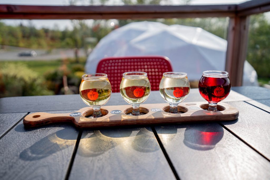 Four round cups hold different ciders, set in a plank of wood on a table. 